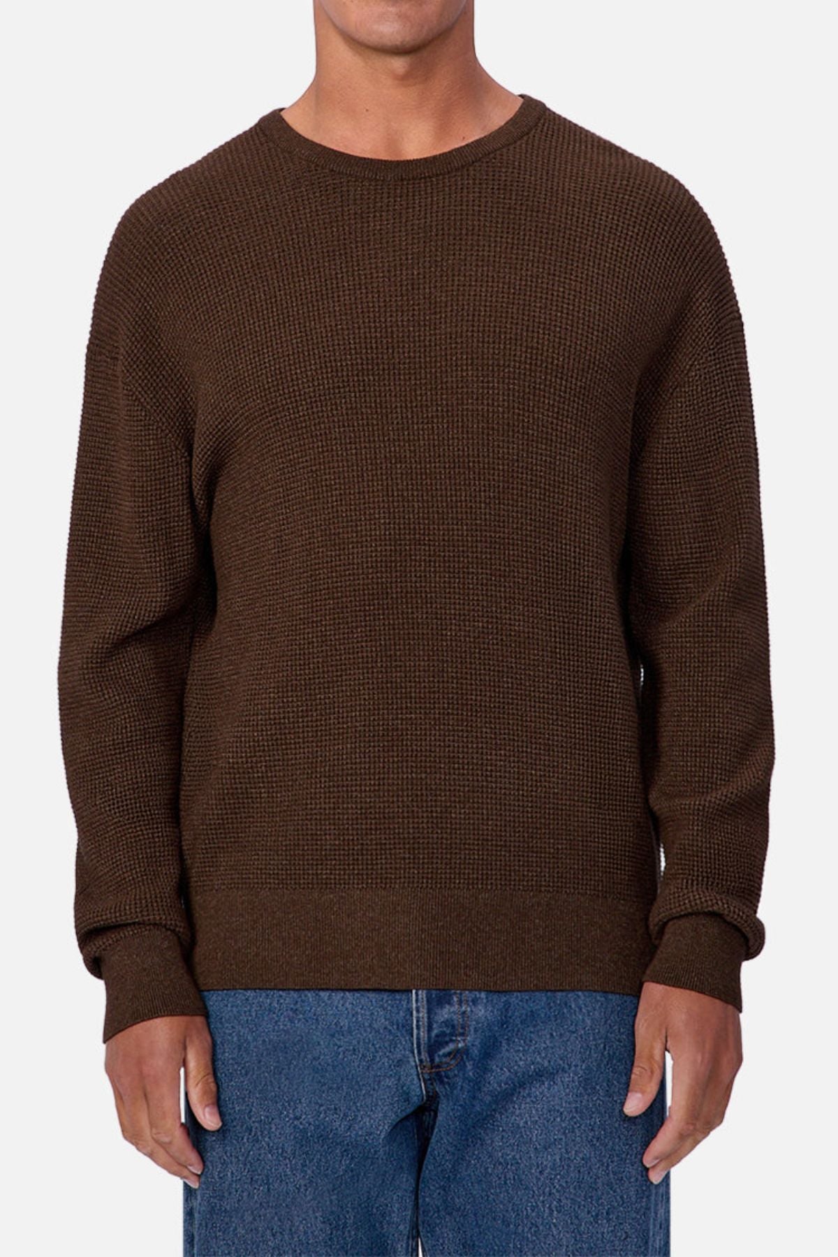 A person wears the Industrie The Philly Waffle Knit, a textured, dark brown men's sweater made from waffle knit fabric, paired with blue jeans. The individual stands against a plain light background with their face not visible.
