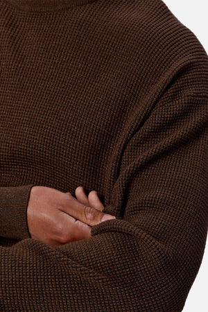 A close-up of a person wearing the Industrie The Philly Waffle Knit, a textured dark brown men's crew neck sweater with a ribbed waffle pattern, arms crossed and hand resting on their upper arm.