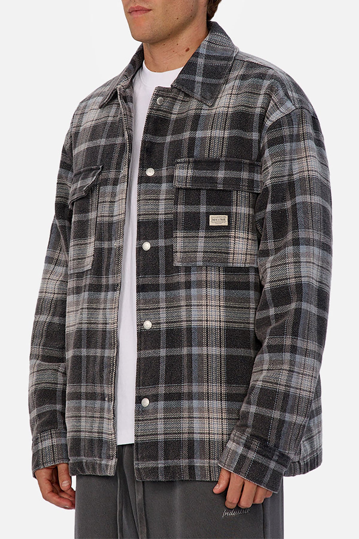 Someone wearing the Industrie The Phoenix Jacket—a gray and black plaid heavyweight shirt jacket—over a white t-shirt and gray pants, standing against a plain white background.