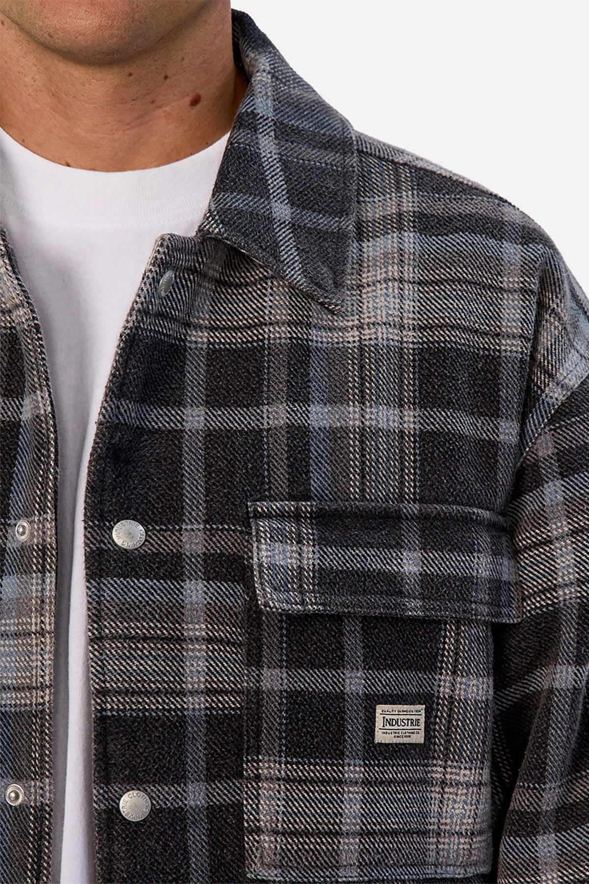 A person wears the Industrie The Phoenix Jacket—a heavyweight black, gray, and blue plaid shirt jacket—over a white t-shirt. A small rectangular "Industrie" label is visible on the front pocket; only the lower half of their face is shown.