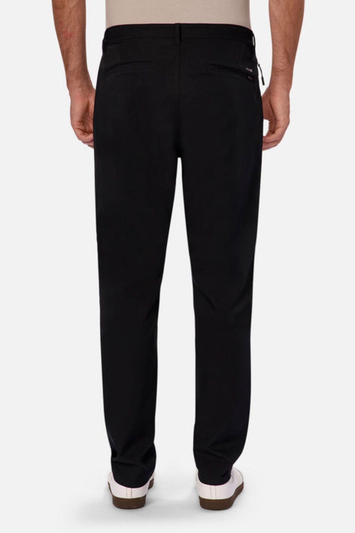 A person is seen from the back wearing black Industrie The Tech Pinehurst Chino Pants, crafted from recycled polyester, paired with white sneakers. They are positioned against a plain background. These flexible trousers feature two rear pockets, and the individual is also sporting a beige top.