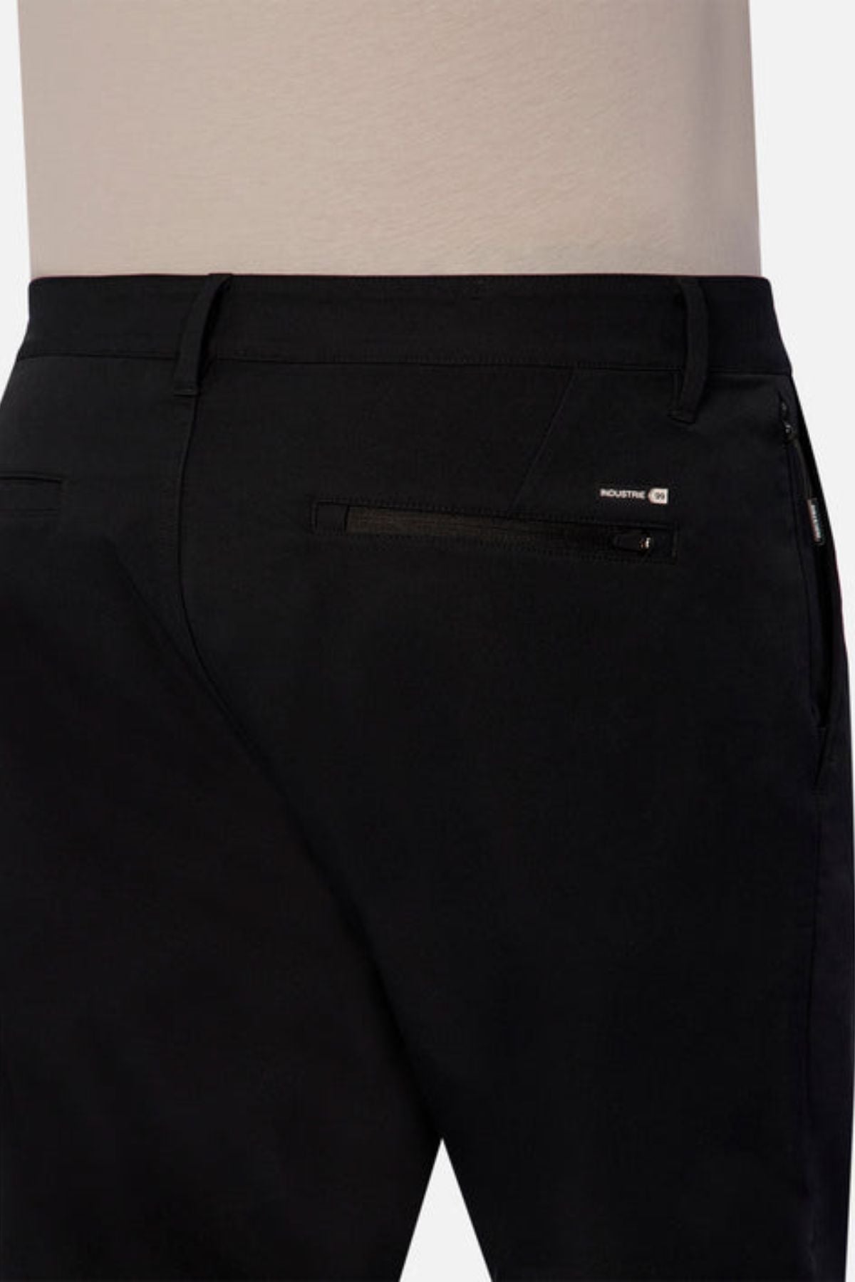 A person wearing black Industrie The Tech Pinehurst Chino Pant, which includes a zipper pocket on the back right side. These pants showcase a subtle logo patch above the pocket and provide exceptional flexibility due to their recycled polyester fabric.