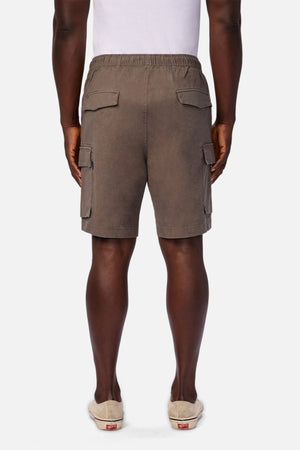 A person stands facing away, wearing a white shirt paired with Industrie The Platoon Combat Short in brown, which features an elastic waistband, side pockets, and back pockets. Beige slip-on shoes with white soles complete the look against a plain white background.