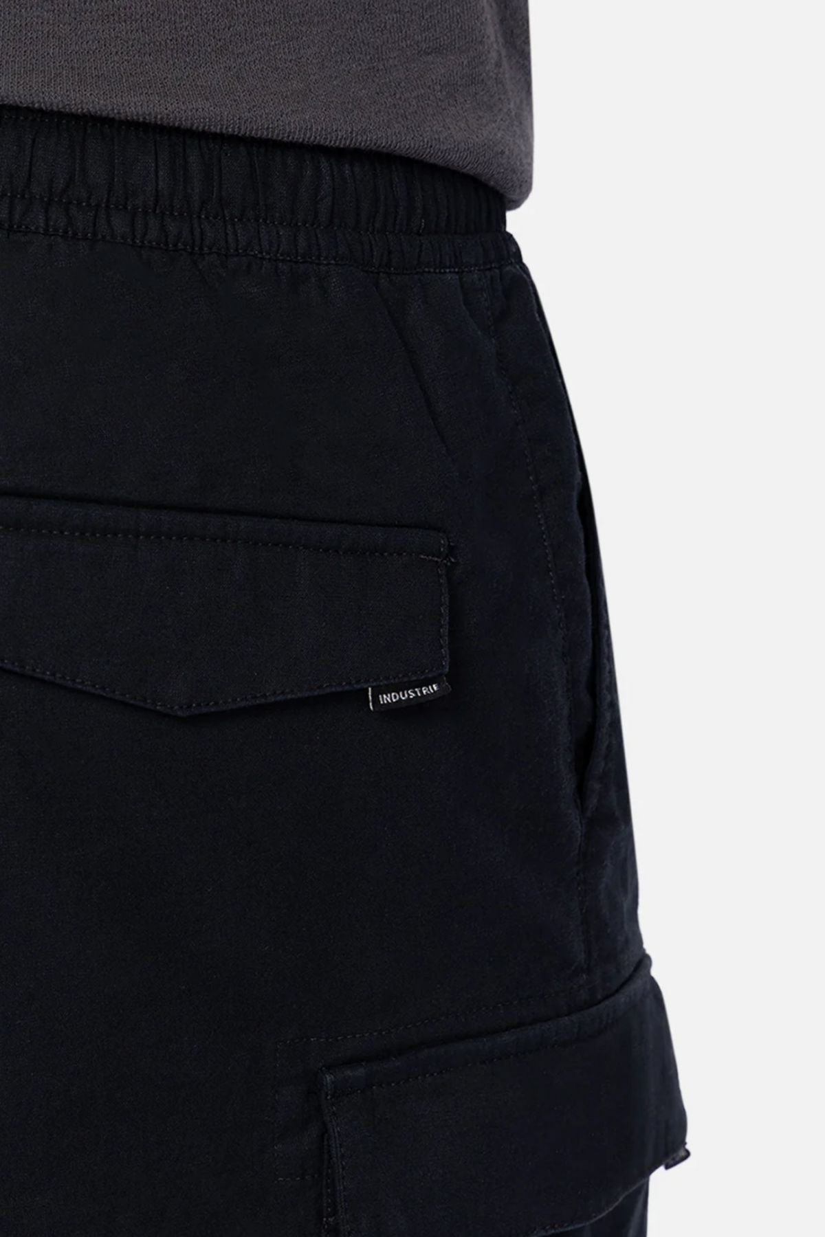 Close-up of a person wearing Industrie The Platoon Combat Short in black, featuring an elastic waistband, flap pockets with a small tag, paired with a tucked-in dark gray shirt.
