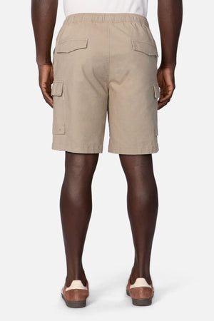 A person is seen from behind wearing Industrie The Platoon Combat Short in light tan with an elastic waistband and four pockets, paired with brown slip-on sandals, standing against a plain white background.