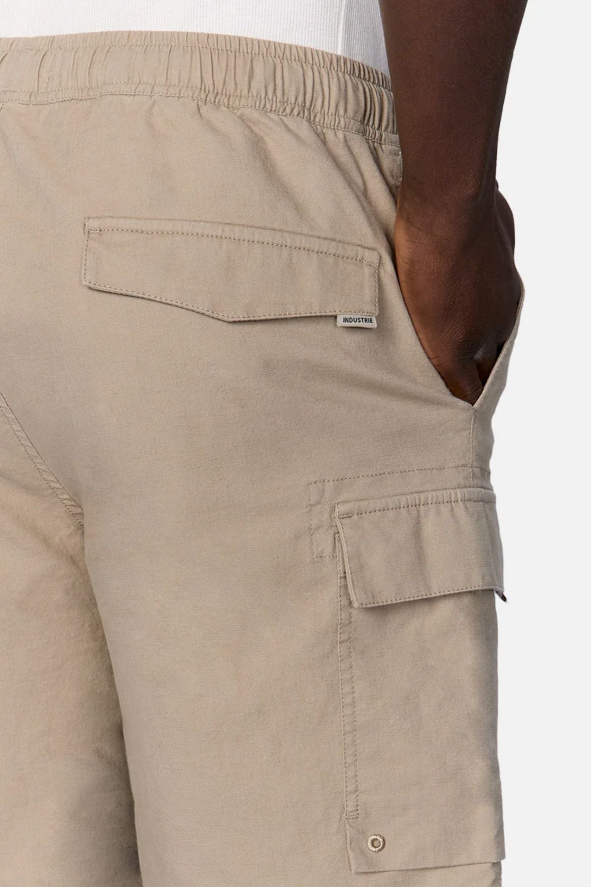 A close-up of a person wearing Industrie The Platoon Combat Short in light beige, featuring a four-pocket design, elastic waistband, flap and back pockets, and a small tag above the back pocket, with one hand in the side pocket.
