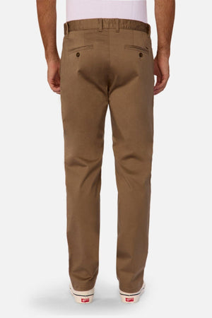 A man is seen from behind wearing Industrie The Regular Cuba Chino Pant in brown with a wider leg fit, belt loops, and two back pockets with button closures, paired with a white shirt and white sneakers.