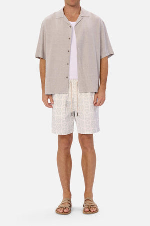 A man wearing a beige short-sleeve button-up shirt over a white T-shirt, paired with Industrie The Saratoga Swim Short featuring a unique print and tan sandals. This casual, summery outfit is perfect for quick-drying convenience at the beach.