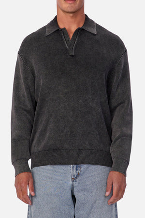 A man wears the Industrie The Sawtelle Knit, a dark gray, long-sleeve zip-collar sweater, paired with light blue jeans and standing against a plain light background—ideal for comfortable everyday wear.