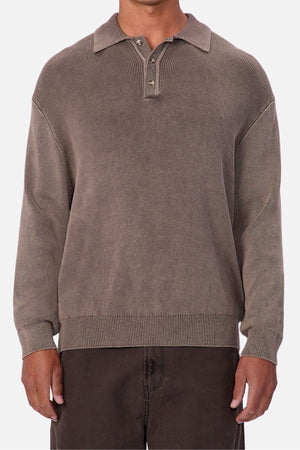 A person wearing the Industrie The Sawtelle Knit—a loose-fitting, long-sleeved polo sweater with a classic collar and three buttons—paired with dark brown pants stands against a plain light background.