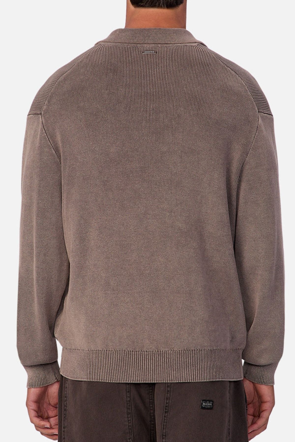 A person is seen from behind in a loose, long-sleeve taupe Industrie The Sawtelle Knit sweater made of 100% cotton with ribbed cuffs and hem, paired with dark pants against a plain neutral background.