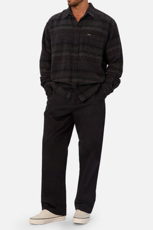 A man stands with his hands in his pockets, wearing the Industrie The Sonoran Long Sleeve Shirt, regular fit black pants, and white slip-on shoes against a plain light background.