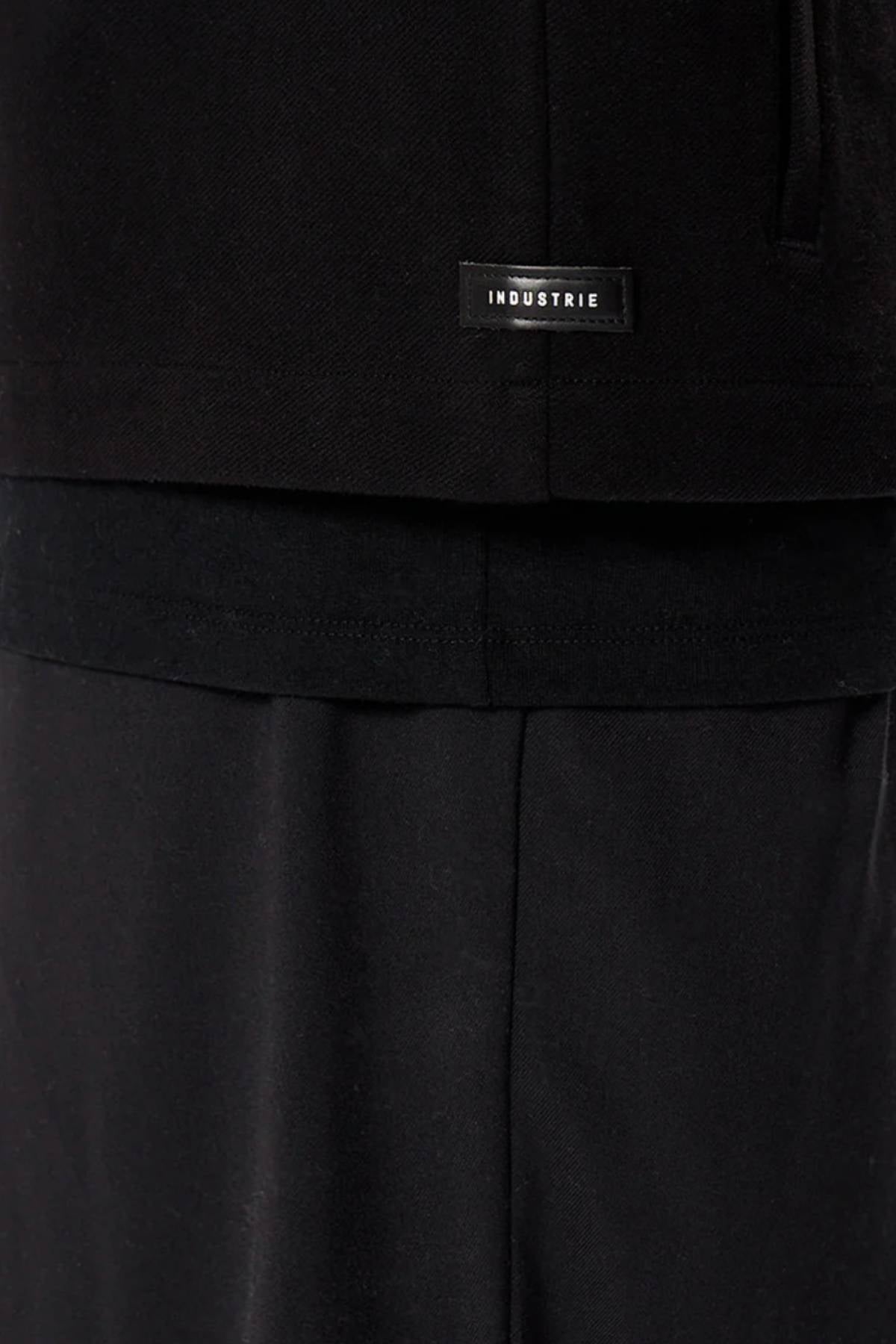 Close-up of a person in black pants and a black shirt layered over another black top, featuring the Industrie logo—a subtle nod to the minimalist design of Industrie The Southport Jacket.