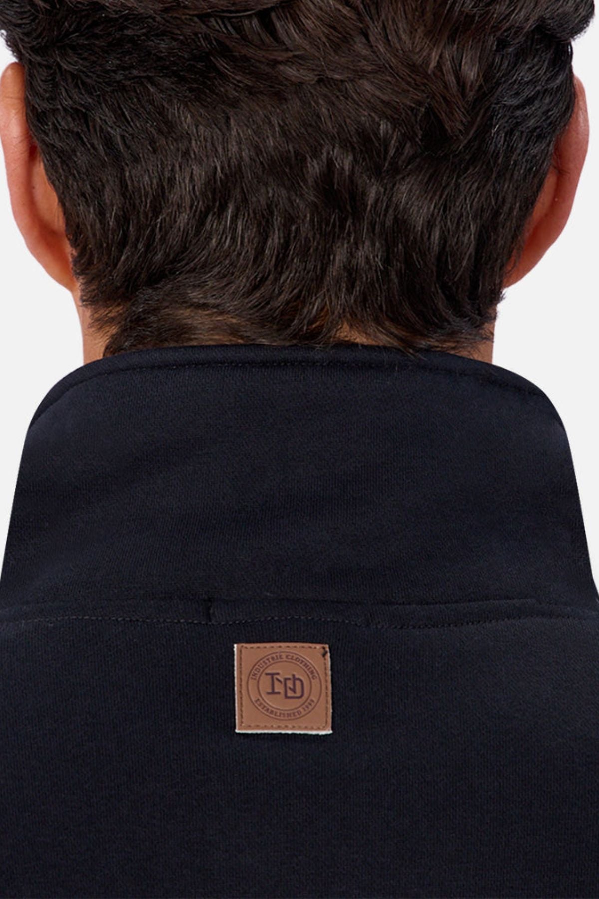 The image shows the back of a man's head and neck with short dark hair and a raised collar. A brown leather patch with a circular logo and text is sewn onto the Industrie The Staunton Track Top men's casual jacket.