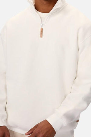 An individual models the Industrie The Staunton Tracktop in off-white, featuring long sleeves, a high collar, branded trims, and a zipper with a tan pull tab. Their face is hidden against a simple, light background.