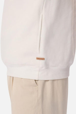 A close-up of a person wearing the Industrie The Staunton Tracktop in cream, featuring branded trims, a side pocket, and a small rectangular tag, paired with light beige pants.