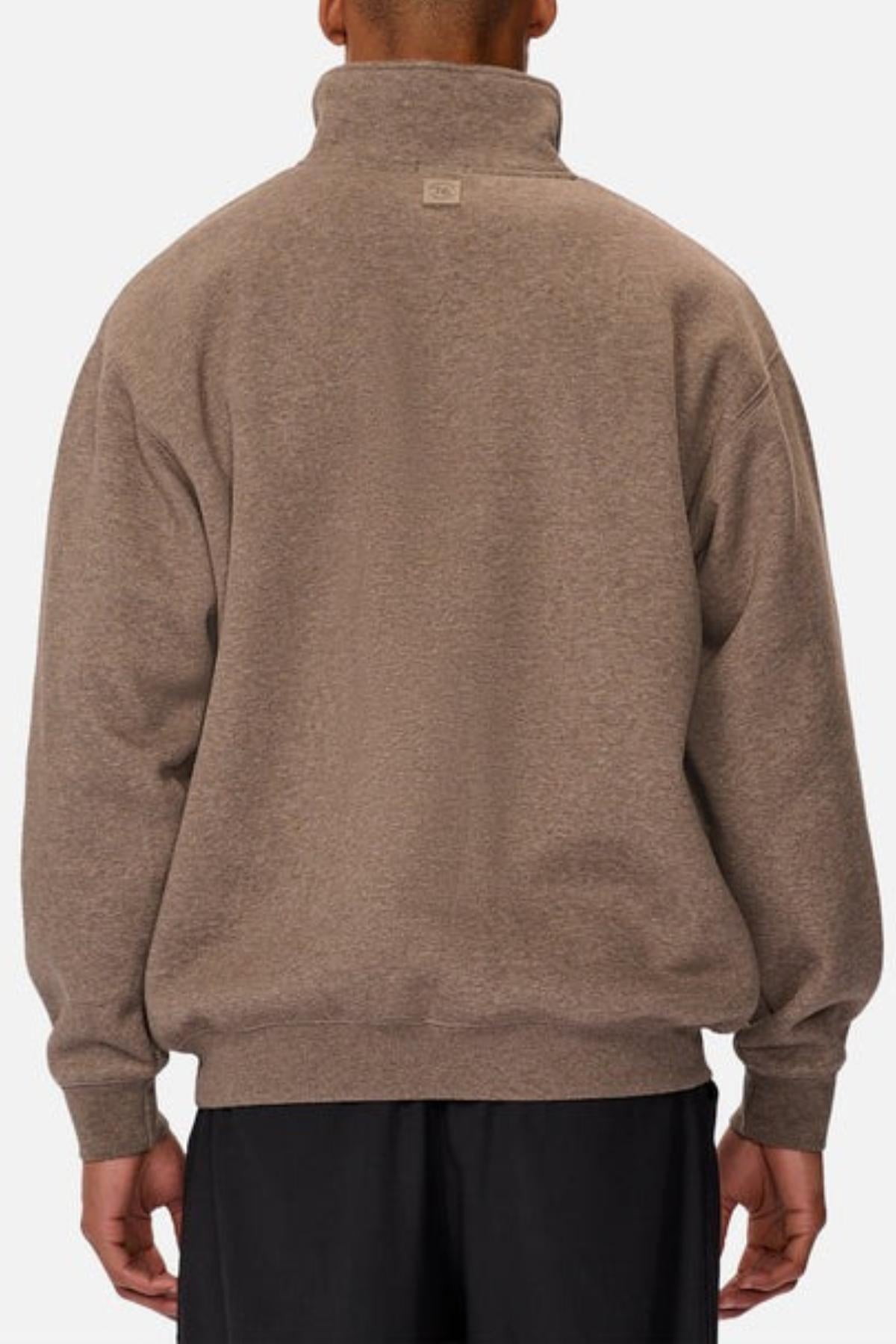 A person is seen from behind wearing the Industrie The Staunton Tracktop, a loose-fit light brown fleece sweatshirt with a high collar and branded trims, paired with black pants against a plain light background.
