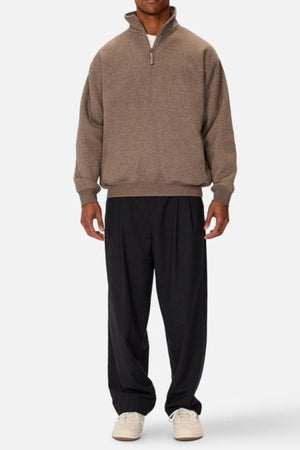 A person faces forward in the Industrie The Staunton Tracktop—a brown quarter-zip with branded trims—paired with loose black pants and white sneakers, set against a plain white background. Their face is not visible.
