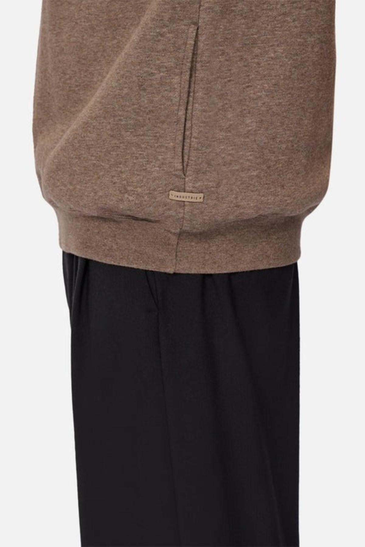 A person models Industrie The Staunton Tracktop in brown, featuring a small label near the hem, paired with loose black pants, shown from the waist down against a plain background.