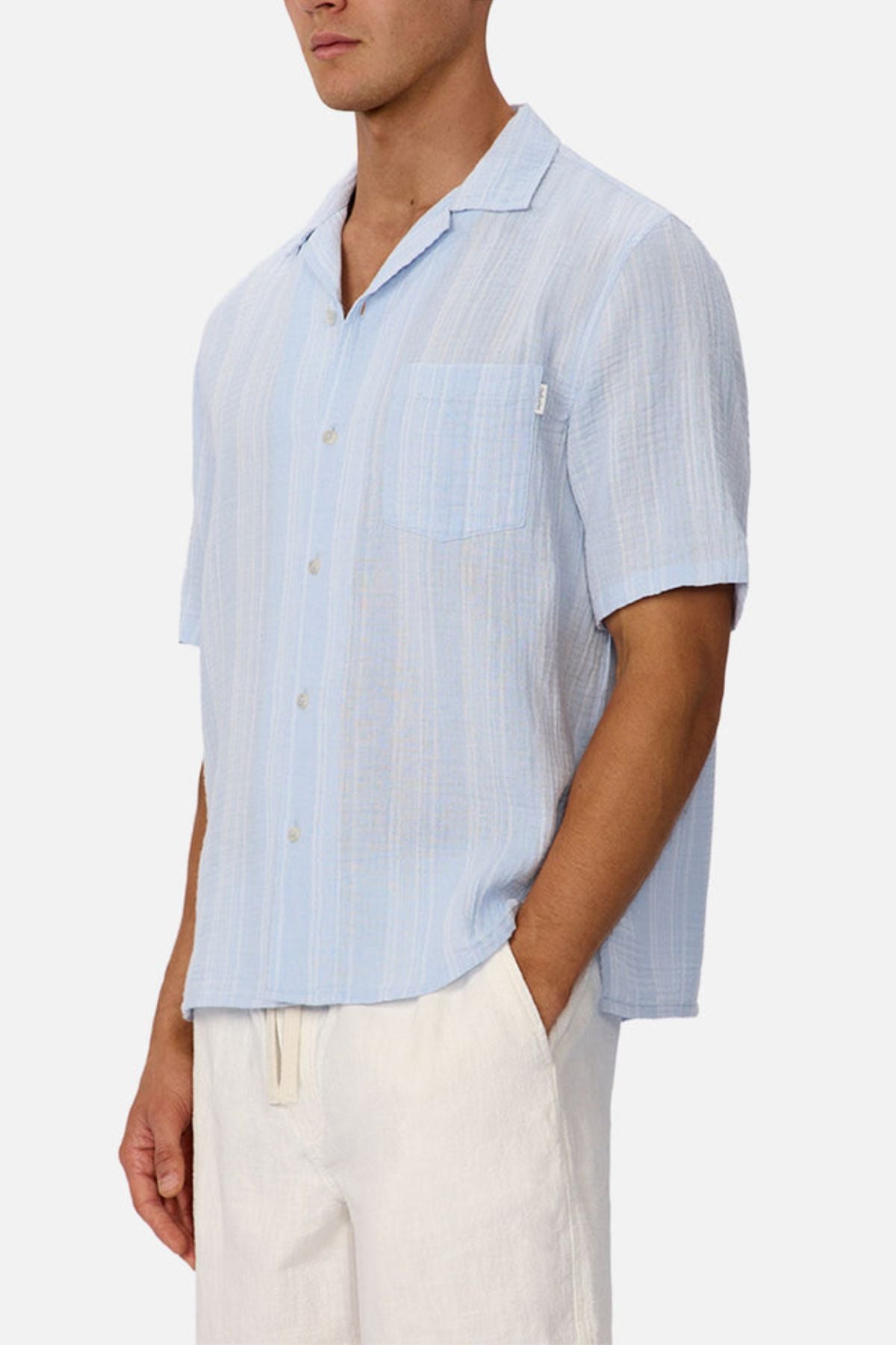 A man wears the Industrie The Striped Seguro Short Sleeve Shirt and white drawstring pants. The shirt is light blue with stripes, short sleeves, a chest pocket, and made from lightweight cotton. The background is plain and neutral.