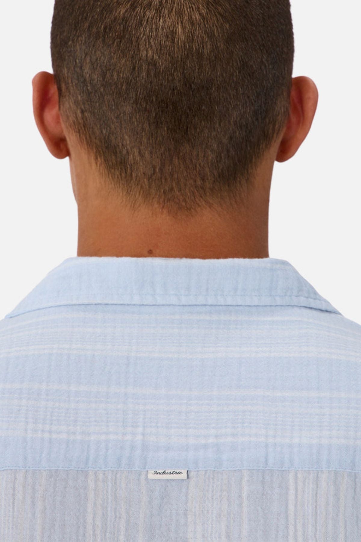 The image shows the back view of a person with short hair wearing the Industrie The Striped Seguro Short Sleeve Shirt in light blue cotton, featuring a small white tag below the collar.