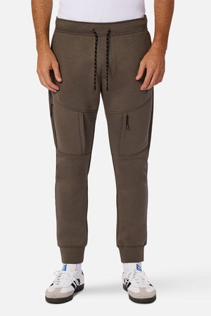 Someone models Industrie The Tech Armoured Track Pant in brown—a jogger-style sweatpant featuring combat pockets and a drawstring waist—paired with white sneakers with blue accents, photographed from the waist down against a plain backdrop.