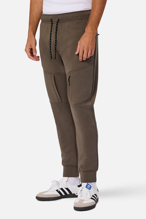 The lower half of a person is shown against a white background, wearing olive green Industrie The Tech Armoured Track Pant joggers with combat pockets and black drawstring, paired with white Adidas sneakers featuring black stripes.