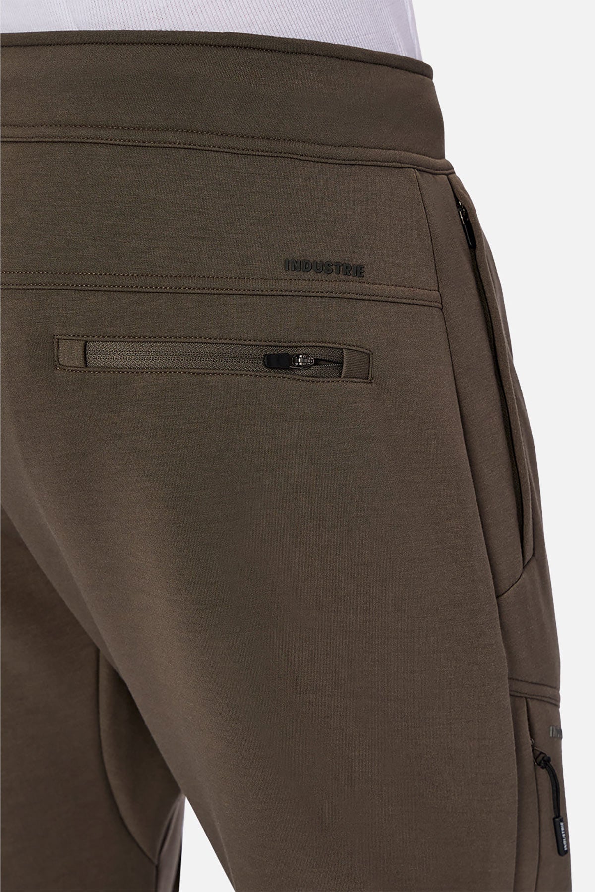 Close-up of a person wearing olive green Industrie The Tech Armoured Track Pant joggers featuring zippered back and combat pockets, with "INDUSTRIE" embroidered above the back pocket.