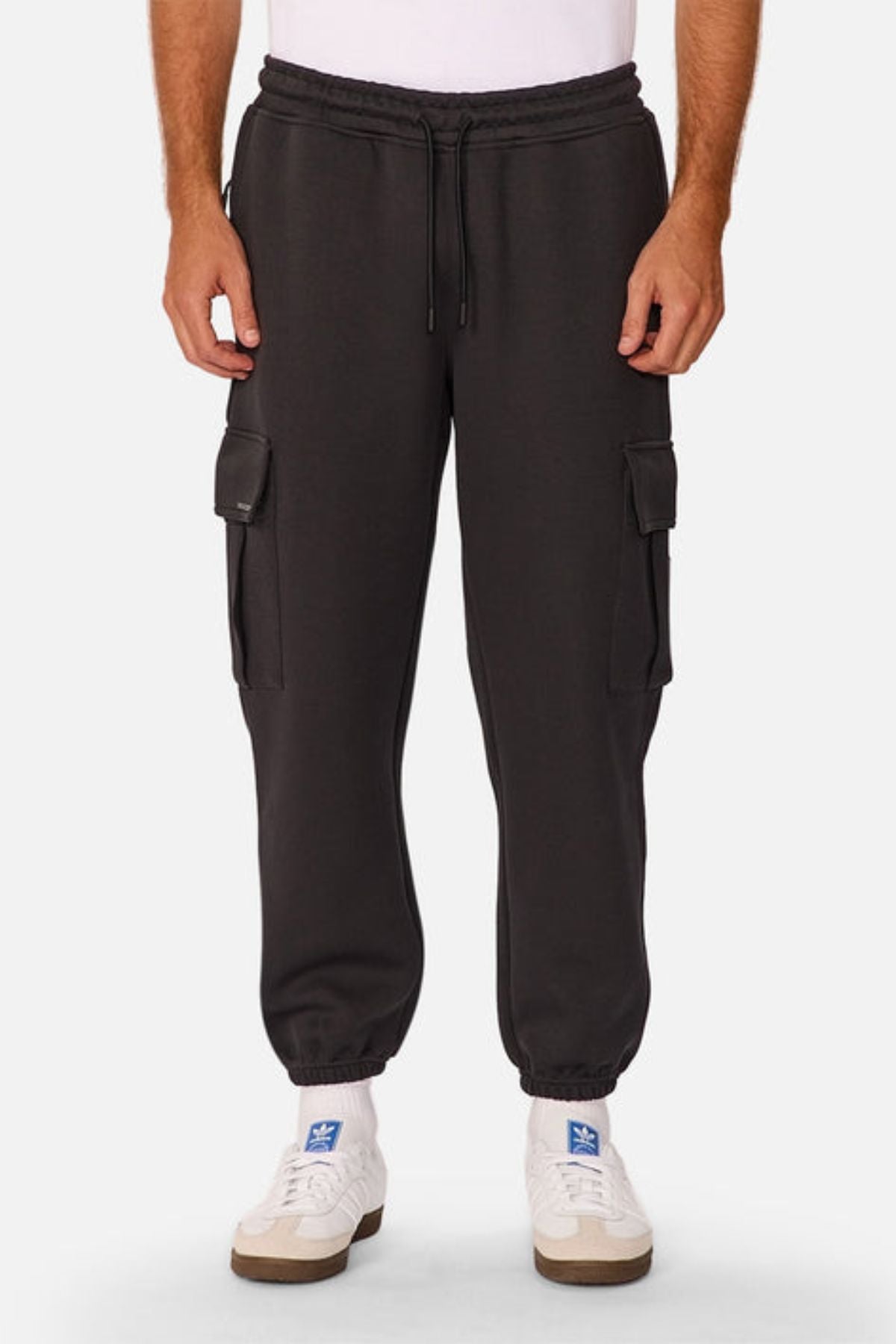 A person wearing Industrie The Tech Combat Trackpant in black with side pockets, white socks, and white sneakers stands against a plain light background. Its customizable fit combines classic style with modern athletic versatility.