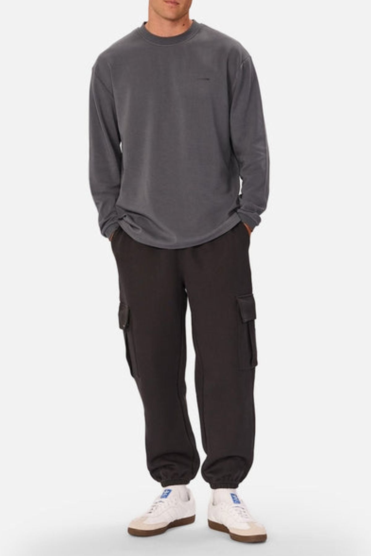 A person wearing a loose gray sweatshirt, Industrie The Tech Combat Trackpant cargo pants with a customizable fit, and white sneakers stands against a plain background with hands in pockets.
