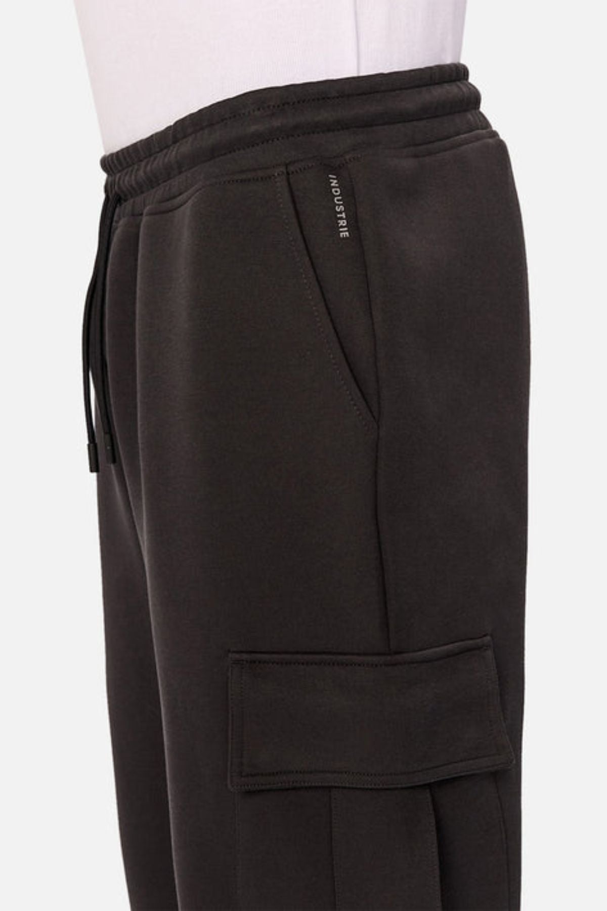 A close-up side view of the Industrie The Tech Combat Trackpant, dark jogger pants with an elastic waistband, drawstring for a customizable fit, side pocket, large flap cargo pocket, and "INDUSTRIE" tag. Perfect for athletic wear.