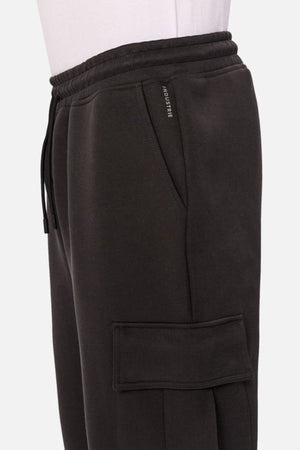 A close-up side view of the Industrie The Tech Combat Trackpant, dark jogger pants with an elastic waistband, drawstring for a customizable fit, side pocket, large flap cargo pocket, and "INDUSTRIE" tag. Perfect for athletic wear.