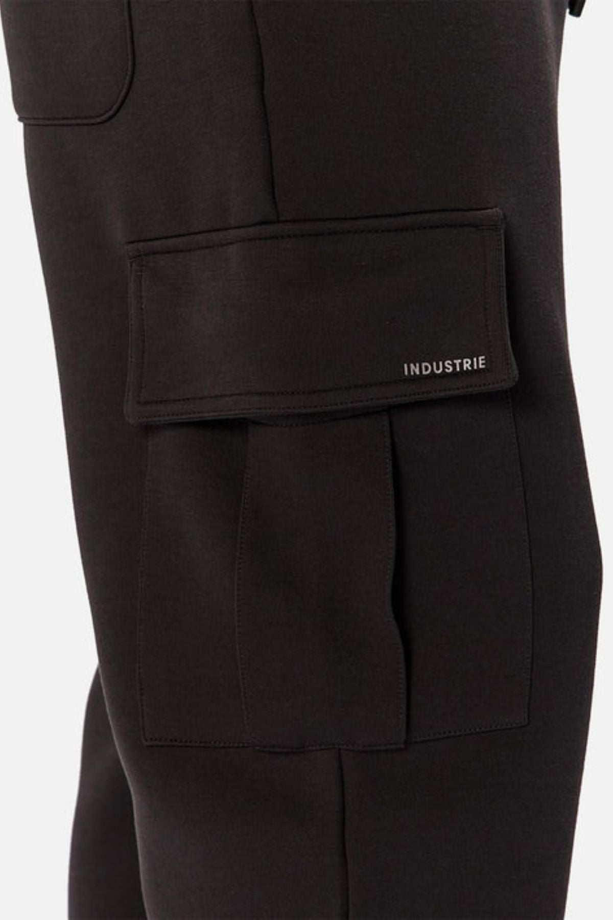 Close-up of Industrie The Tech Combat Trackpant in black, showing a leg with a large flap pocket featuring "INDUSTRIE" in small white letters. The athletic design offers a customizable fit, set against a plain light background.