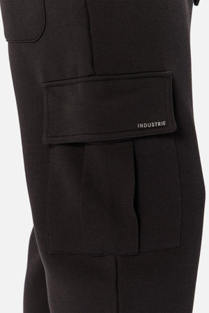 Close-up of Industrie The Tech Combat Trackpant in black, showing a leg with a large flap pocket featuring "INDUSTRIE" in small white letters. The athletic design offers a customizable fit, set against a plain light background.