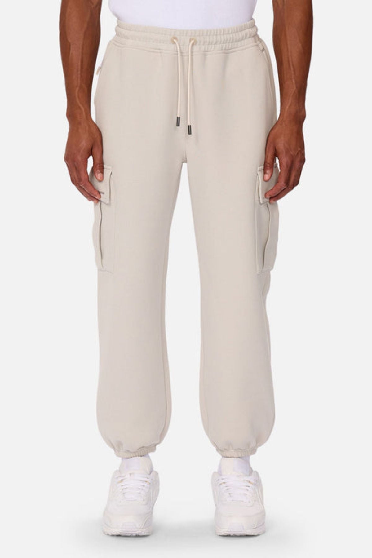 A person wears Industrie The Tech Combat Trackpant in light beige with elastic cuffs and side pockets, styled with a white shirt, socks, and sneakers for an athletic look combining comfort and performance against a plain background.