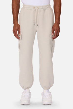 A person wears Industrie The Tech Combat Trackpant in light beige with elastic cuffs and side pockets, styled with a white shirt, socks, and sneakers for an athletic look combining comfort and performance against a plain background.