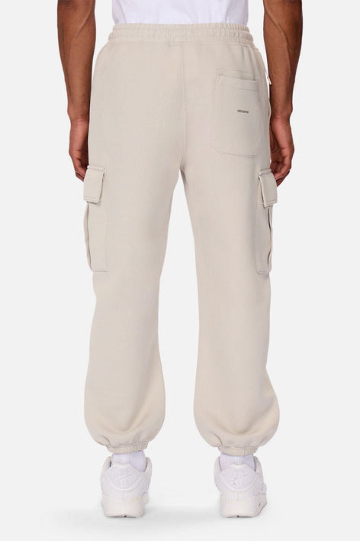 A person is seen from behind wearing Industrie The Tech Combat Trackpant in light beige with elastic cuffs, rear and side flap pockets, styled with white sneakers and a white T-shirt for a modern athletic look.