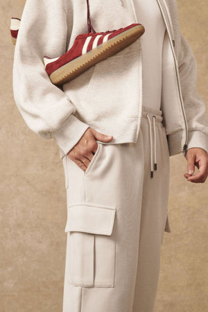 Wearing Industrie The Tech Combat Trackpant in light beige, a matching zip-up hoodie, and a white shirt, the person poses with one hand in their pocket while carrying a red Adidas sneaker with white stripes over their shoulder.