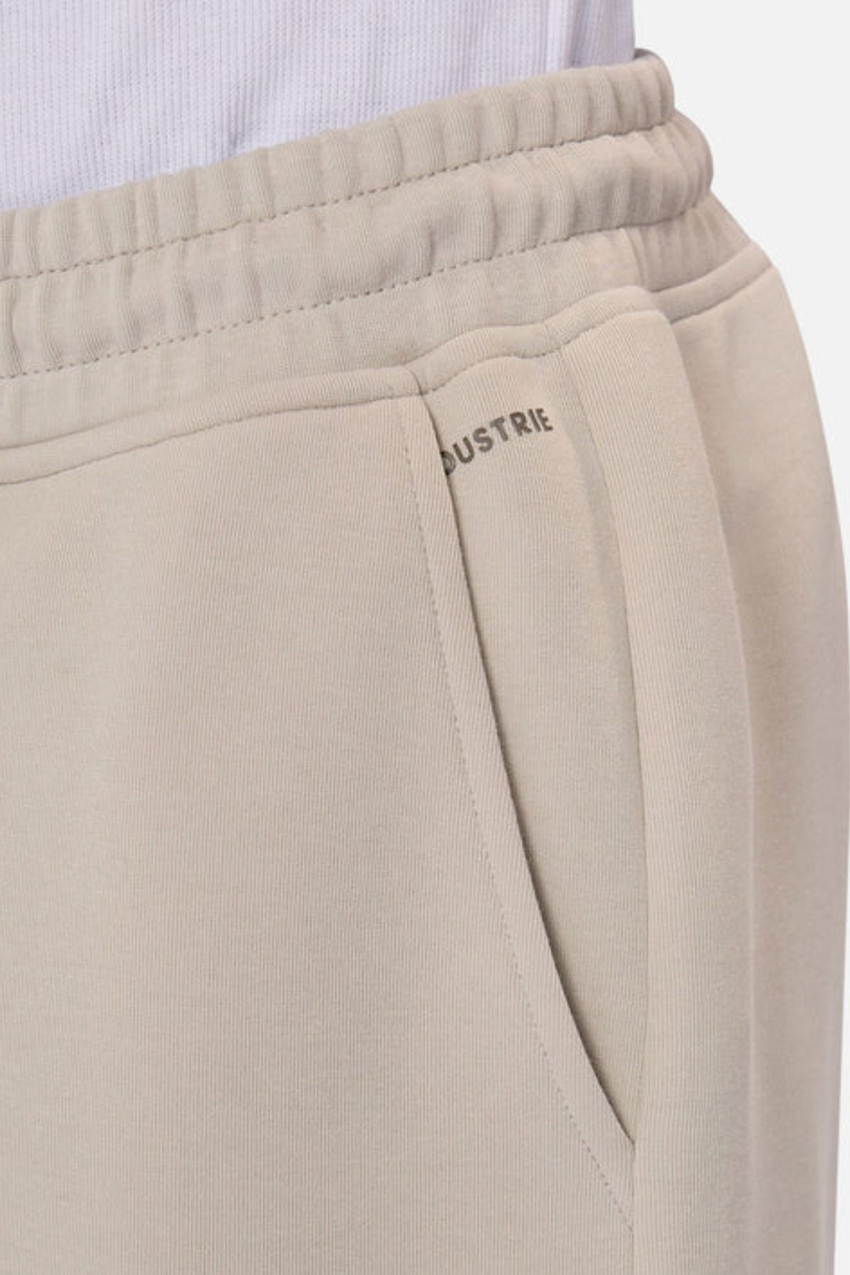 Close-up of someone in Industrie The Tech Combat Trackpant, light beige with elastic waistband and side pocket. The "INDUSTRIE" logo is partly visible near the pocket, styled with a white shirt for an effortless athletic look.