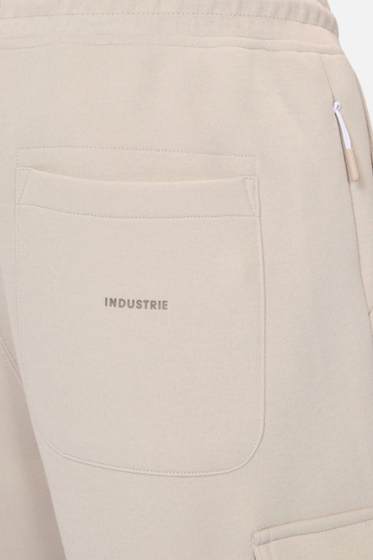 Close-up of the back pocket on Industrie The Tech Combat Trackpant in beige, with "INDUSTRIE" printed above the pocket and a white zipper pull by the side seam, showcasing its modern athletic performance style.