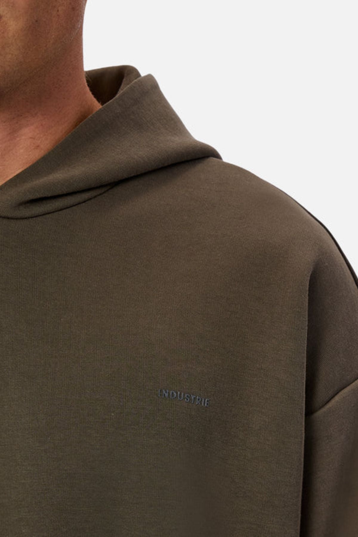 Close-up of a person in Industrie The Tech Del Sur Hood, olive-green with a small embroidered "INDUSTRIE" logo on the chest. Made with advanced athletic fabric; only part of the face, neck, and shoulder are shown.