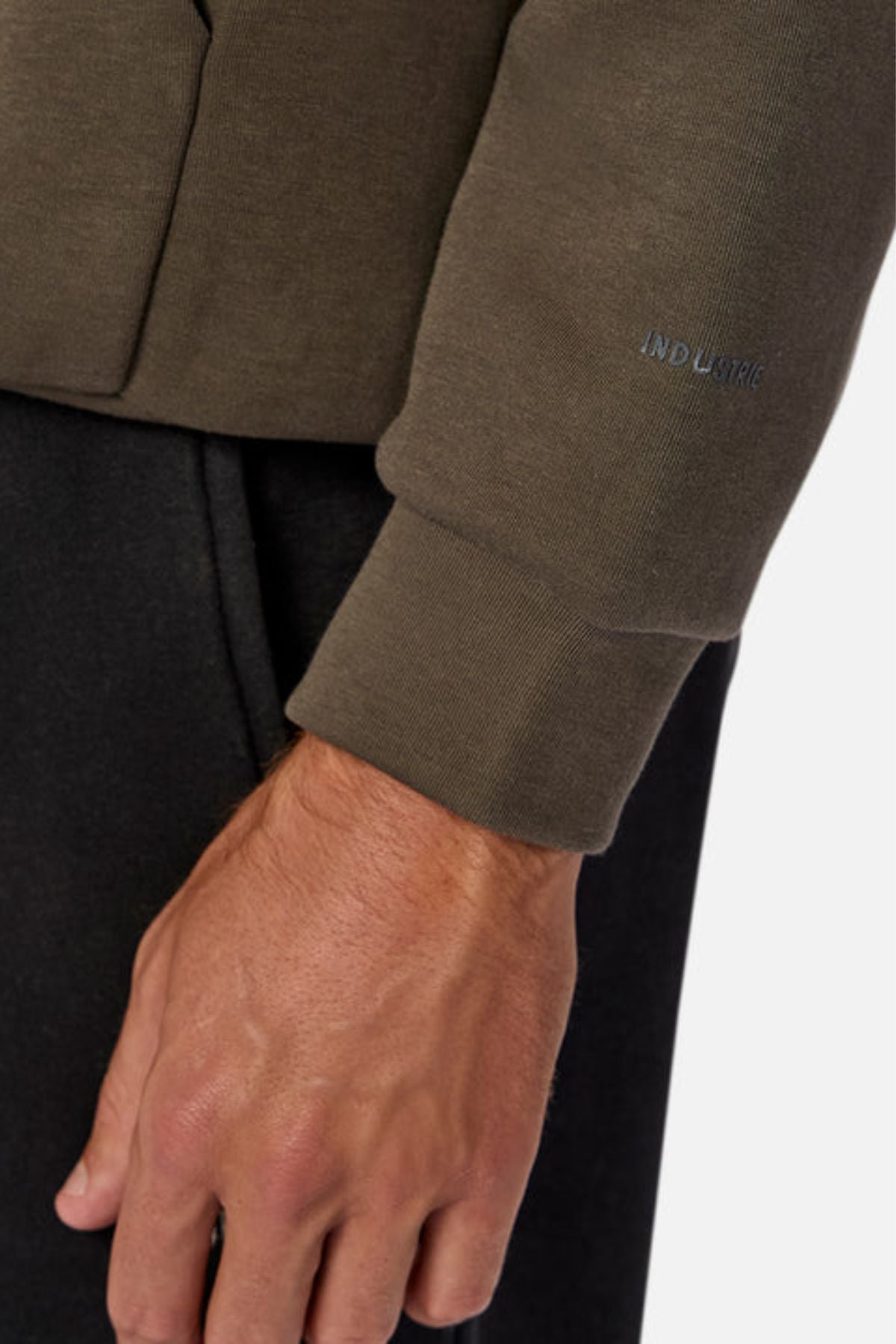 A close-up shows a person’s arm in the Industrie The Tech Del Sur Hood, featuring advanced athletic fabric in olive green with "INDUSTRIC" embroidered near the cuff; black pants appear in the background.