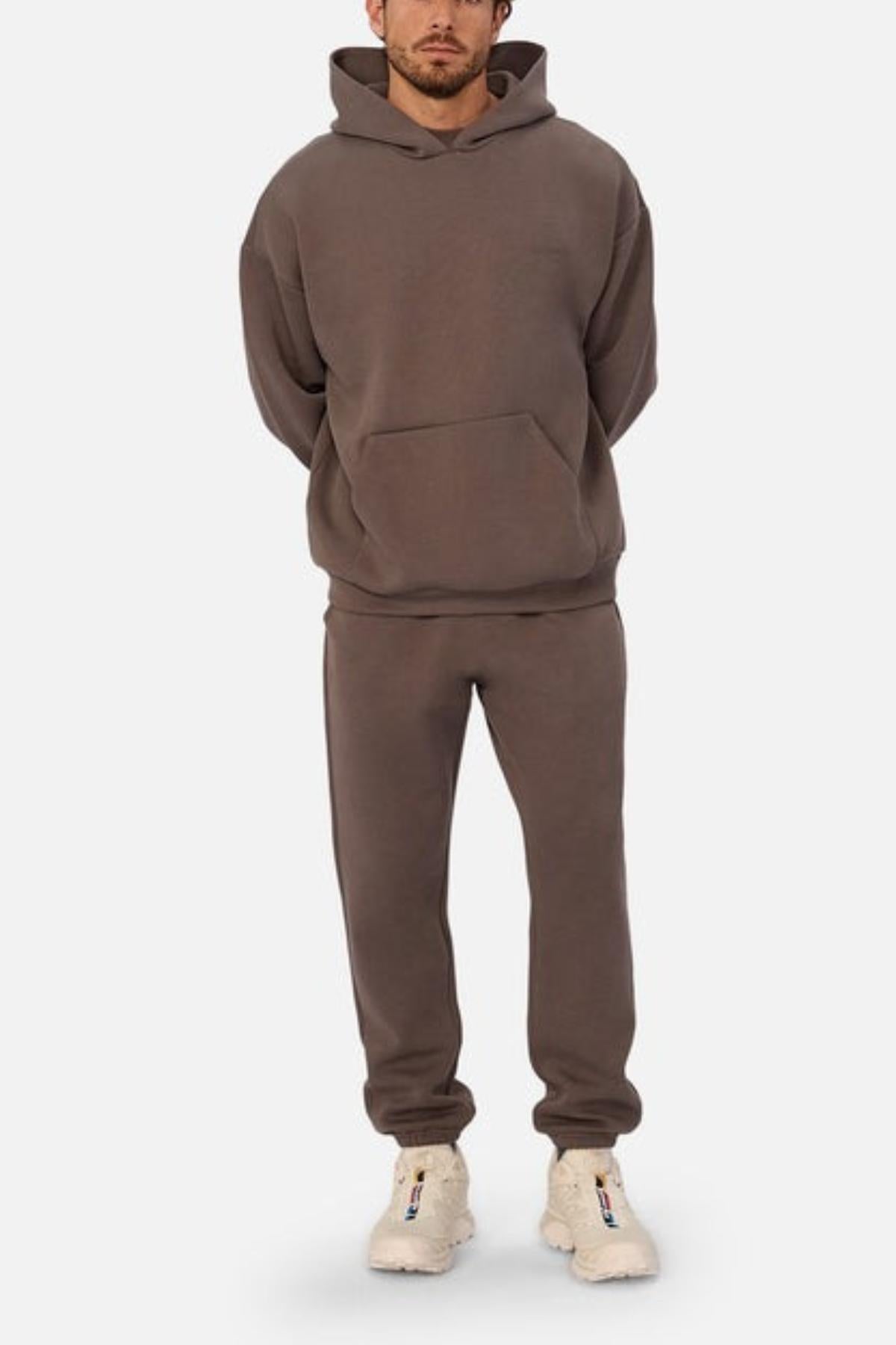 A man wears the Industrie The Tech Del Sur Hood in loose-fitting brown with matching joggers and off-white sneakers, standing hands behind his back against a plain, light background.
