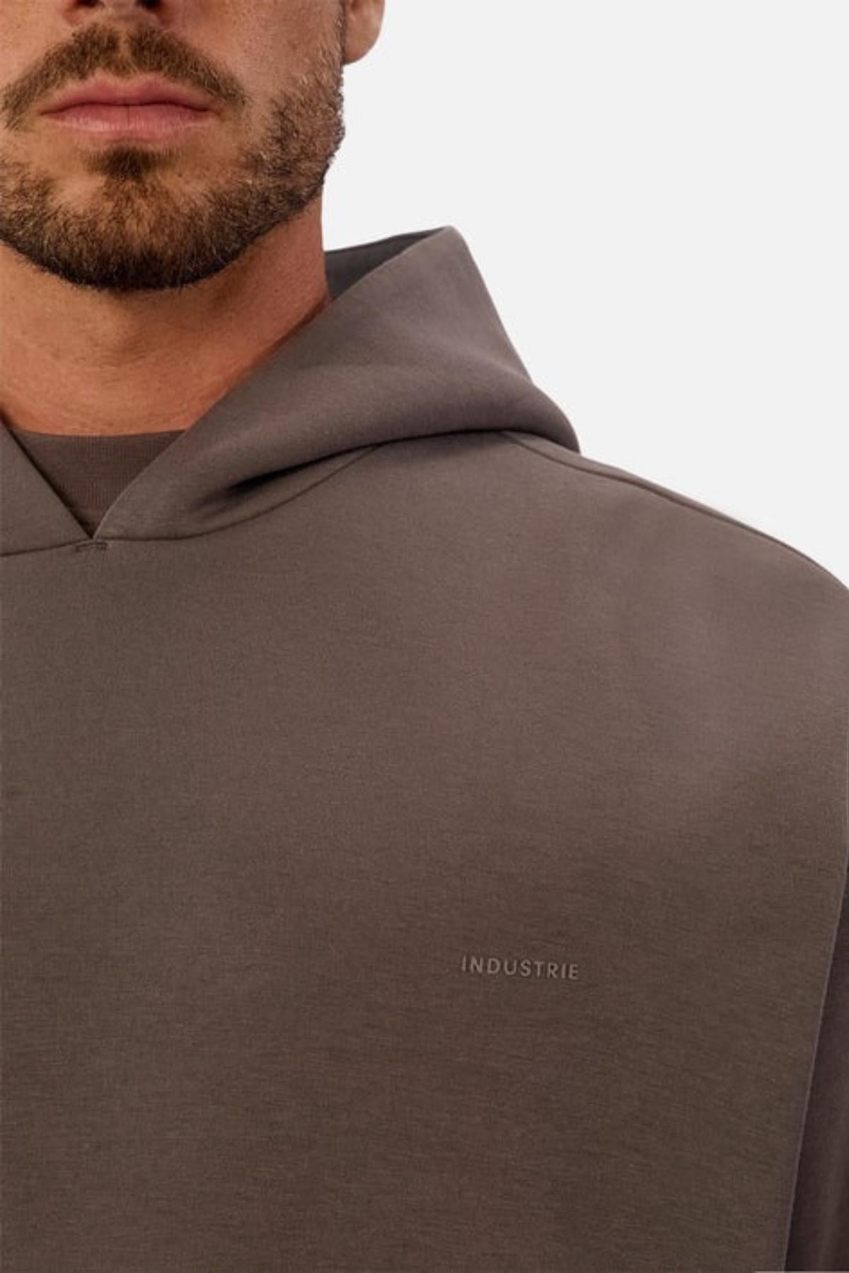 A close-up of a man in the Industrie The Tech Del Sur Hood in taupe, featuring a subtly embroidered INDUSTRIE logo on the chest. His face is partly visible with a short beard and mustache against a plain white background.
