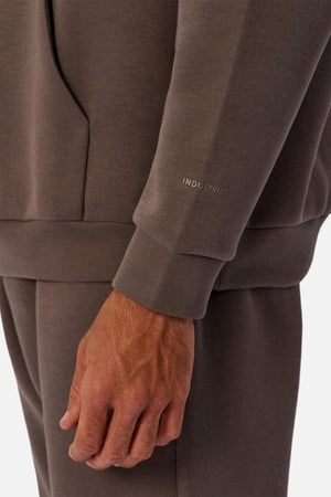 A close-up of a person in the Industrie The Tech Del Sur Hood, a brown men's pullover with matching pants. Their left hand is by their side, and “INDUSTRIE” is printed near the wrist on the sleeve.