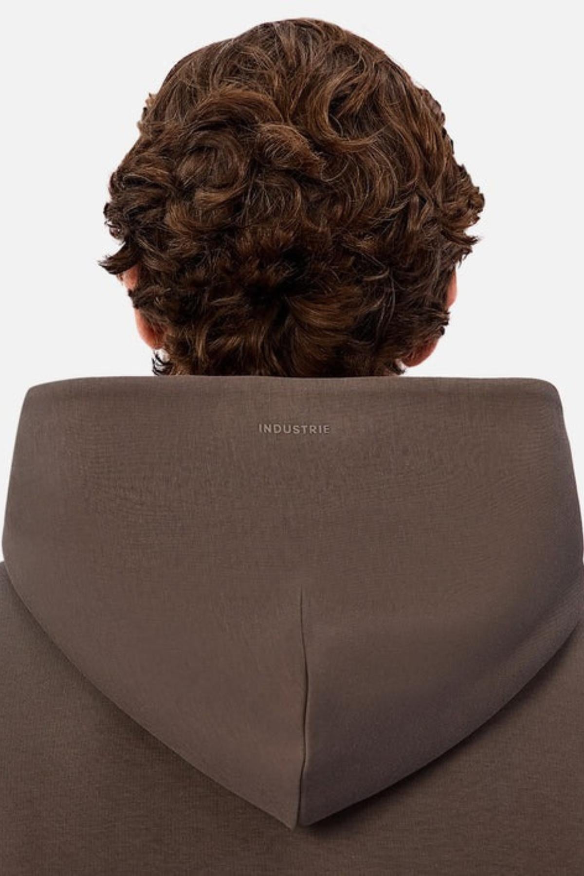 A person with curly brown hair is shown from behind, wearing the Industrie The Tech Del Sur Hood in brown with a small white INDUSTRIE logo on the back of the hood. The plain white background highlights its advanced athletic fabrication.