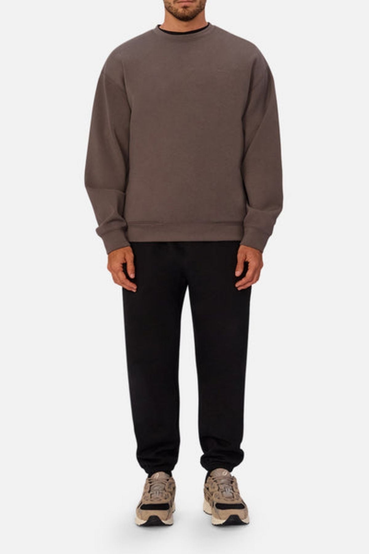 A man stands facing forward in modern athleisure, wearing a loose-fitting Industrie The Tech Del Sur Sweat, black 4-way stretch sweatpants, and beige chunky sneakers against a plain light gray background.