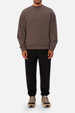 A man stands facing forward in modern athleisure, wearing a loose-fitting Industrie The Tech Del Sur Sweat, black 4-way stretch sweatpants, and beige chunky sneakers against a plain light gray background.