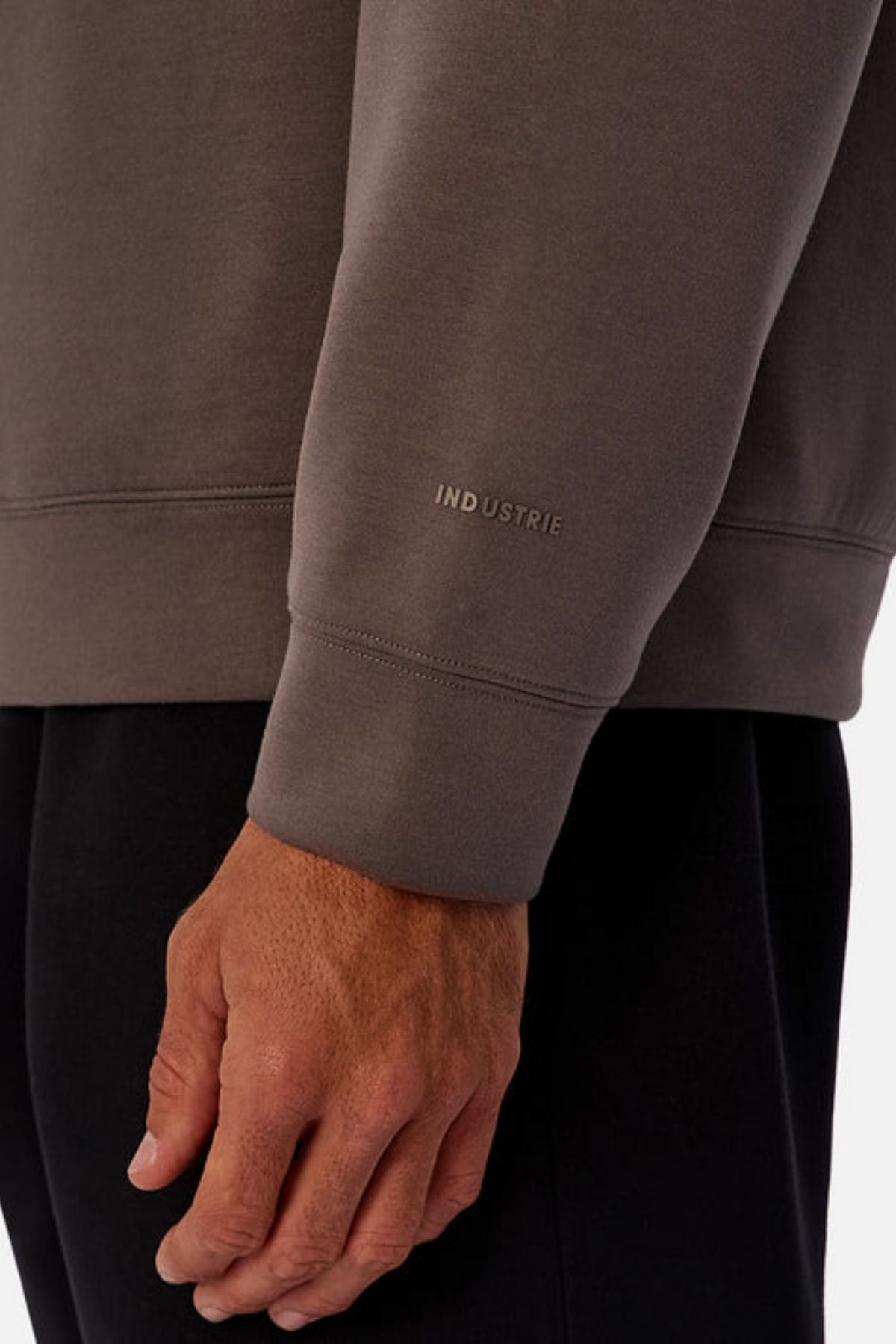 A close-up of a left arm wearing the Industrie The Tech Del Sur Sweat, a taupe athleisure sweatshirt with "INDUSTRIE" near the cuff, paired with black pants. The relaxed hand rests at the person's side.