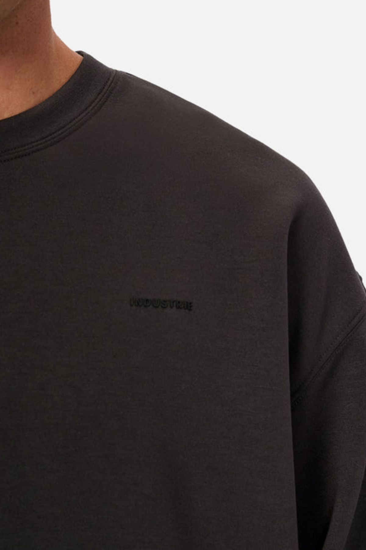 Close-up of a person in the Industrie The Tech Del Sur Sweat—a plain black crewneck sweatshirt with subtle "INDUSTRIE" chest embroidery. Only the neck, shoulder, and upper chest are shown, highlighting this modern athleisure essential.